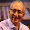 Smiling older man with glasses, wearing a light checkered shirt, in front of a bookshelf backdrop.