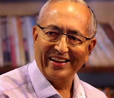 Smiling older man with glasses, wearing a light checkered shirt, in front of a bookshelf backdrop.