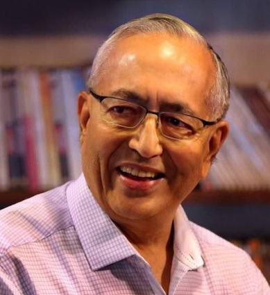 Smiling older man with glasses, wearing a light checkered shirt, in front of a bookshelf backdrop.