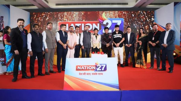 Group of formally dressed people standing on a stage behind a podium with 'NATION 27' branding and sparkly backdrop.