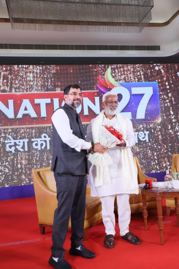 nation27_2 | No. 1 Indian Media News Portal Two men on a stage exchange a bouquet, with a colorful banner reading 'NATION 27' behind them on a red-carpeted set.