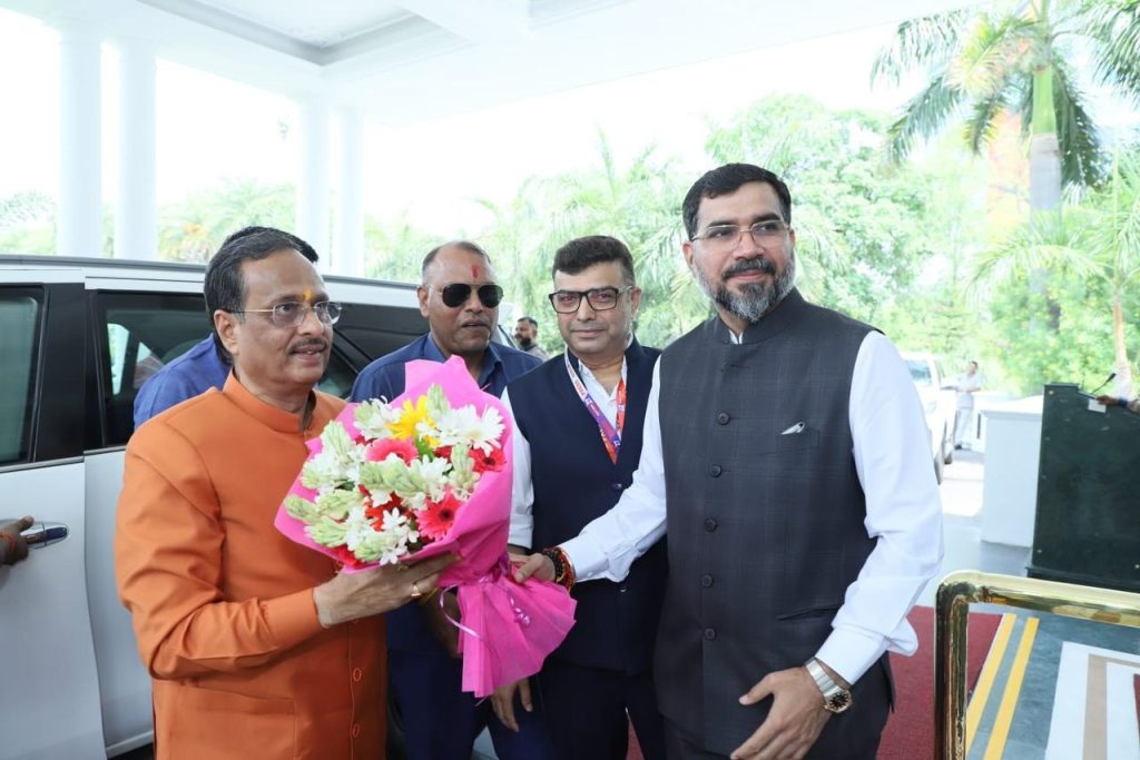 nation27_3 | No. 1 Indian Media News Portal Man in an orange kurta presents a colorful bouquet to a man in a dark waistcoat, as two other men stand nearby outdoors near a white car with greenery in the background.