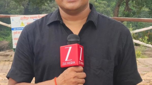 A male reporter in a black shirt holds a microphone labeled 'ZEE MEDIA' outdoors with trees in the background.