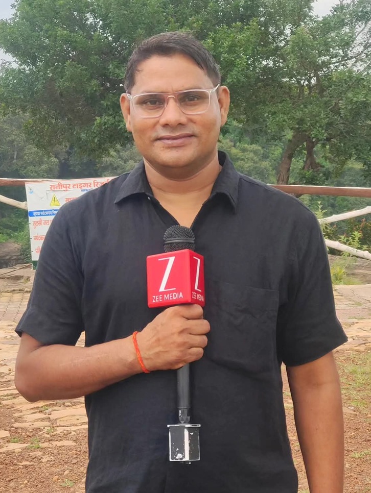 A male reporter in a black shirt holds a microphone labeled 'ZEE MEDIA' outdoors with trees in the background.