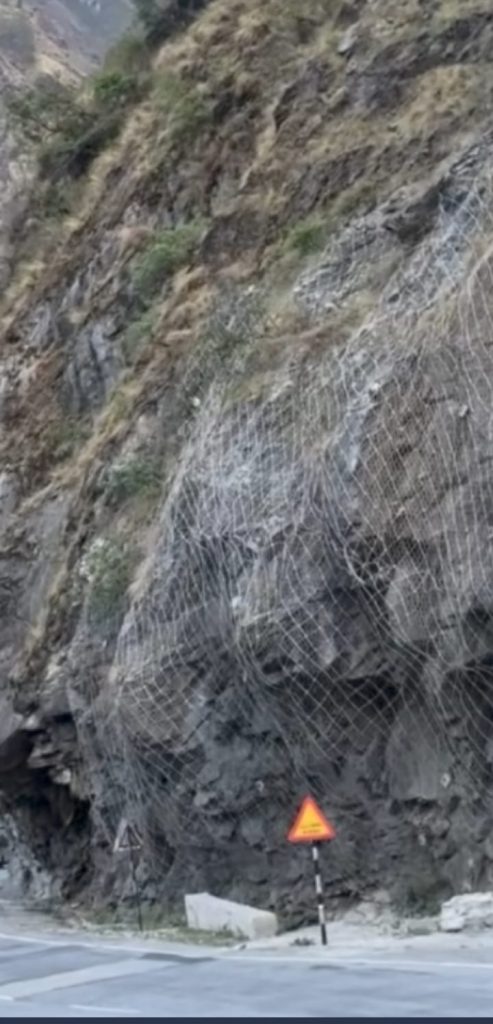 pahad | No. 1 Indian Media News Portal Steep rocky hillside reinforced with white safety netting beside a road; orange warning sign on a striped post.
