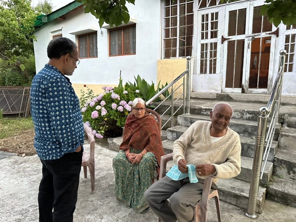 pahad_shrivastava | No. 1 Indian Media News Portal Man in a blue patterned shirt stands and talks to two elderly people seated in plastic chairs outside a white house with steps and railing, a garden with pink hydrangeas nearby.