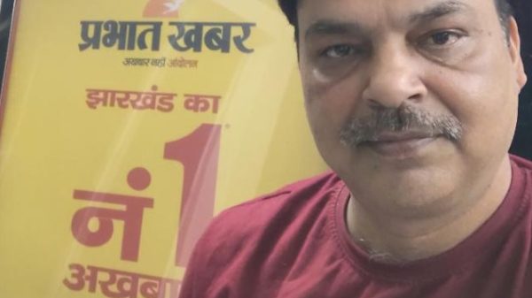 Selfie of a man with a maroon shirt standing in front of a yellow Prabhat Khabar newspaper poster advertising Jharkhand's number 1 newspaper.
