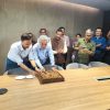 Group of colleagues gathered around a conference table as two men slice a large chocolate cake while others watch and smile in a modern meeting room.