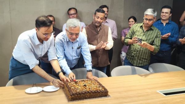 Group of colleagues gathered around a conference table as two men slice a large chocolate cake while others watch and smile in a modern meeting room.