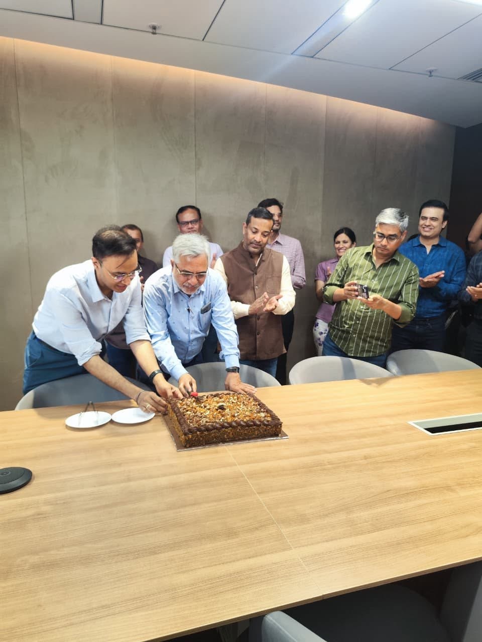 Group of colleagues gathered around a conference table as two men slice a large chocolate cake while others watch and smile in a modern meeting room.