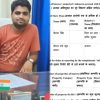 Man in an orange t-shirt sits at a desk with an Indian flag on a document holder in front of a blue curtain backdrop.