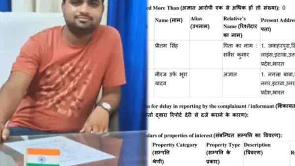 Man in an orange t-shirt sits at a desk with an Indian flag on a document holder in front of a blue curtain backdrop.