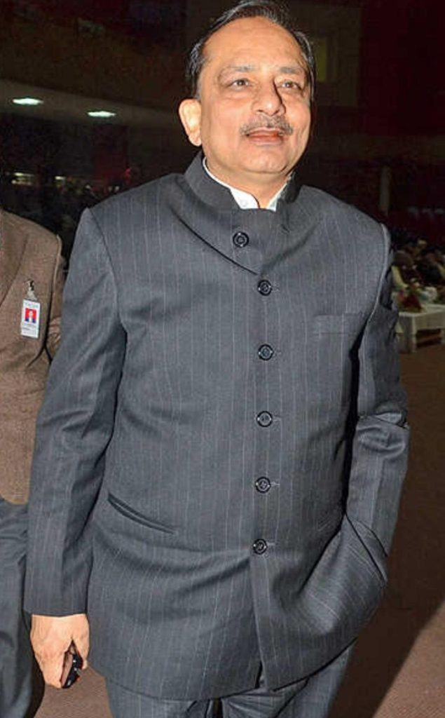 Man wearing a grey pinstriped suit with a stand-up collar at a formal indoor event, hands by his sides.