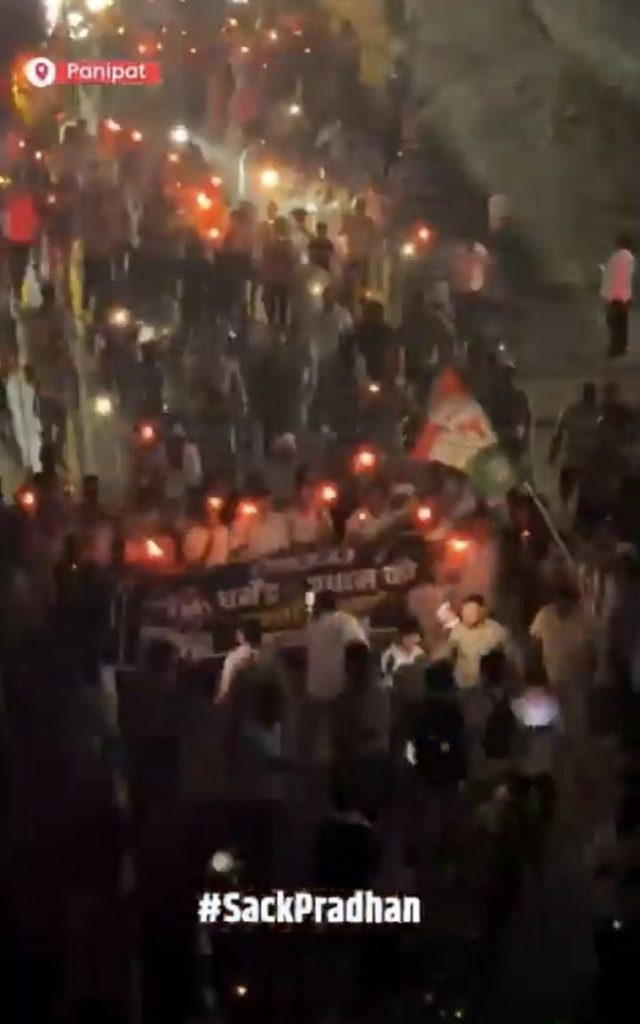 Screenshot | No. 1 Indian Media News Portal Night-time protest: dense crowd marching on a street with torches, banners, and a visible 'Sack Pradhan' banner (Panipat watermark).