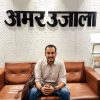 Man sitting on a brown leather couch in an office lobby, with a patterned wall and the Hindi sign 'Amar Ujala' behind him; a brown bag rests beside him.