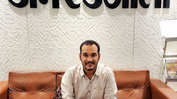 Man sitting on a brown leather couch in an office lobby, with a patterned wall and the Hindi sign 'Amar Ujala' behind him; a brown bag rests beside him.