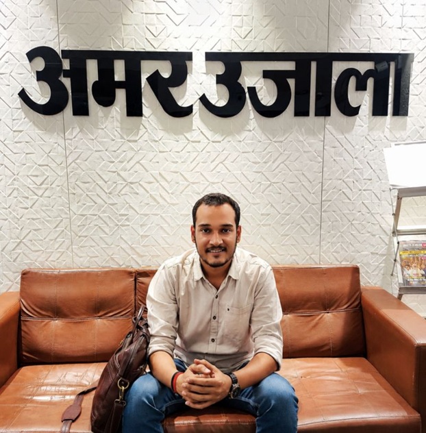 Man sitting on a brown leather couch in an office lobby, with a patterned wall and the Hindi sign 'Amar Ujala' behind him; a brown bag rests beside him.
