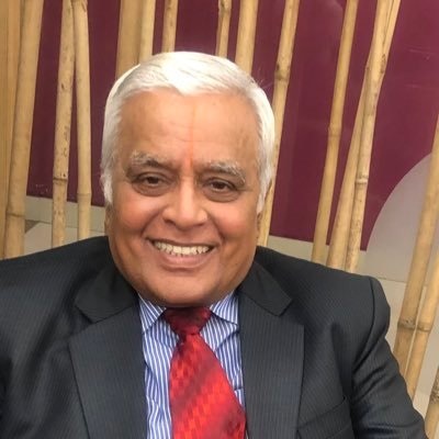 Smiling older man with white hair in a dark suit and red tie, seated indoors with bamboo decor in the background.
