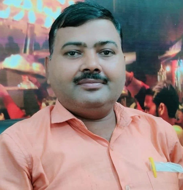 Portrait of a man with a short black mustache wearing a peach shirt, seated indoors with a colorful background behind him.