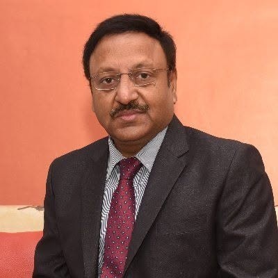rajeev kumar | No. 1 Indian Media News Portal Professional portrait of a man in a dark suit and red tie with glasses against an orange backdrop.