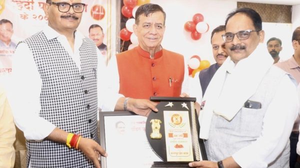 Three men in traditional Indian attire at an award ceremony; the man in the center hands a framed trophy to the man on the right as others look on, with red and white balloons in the background.