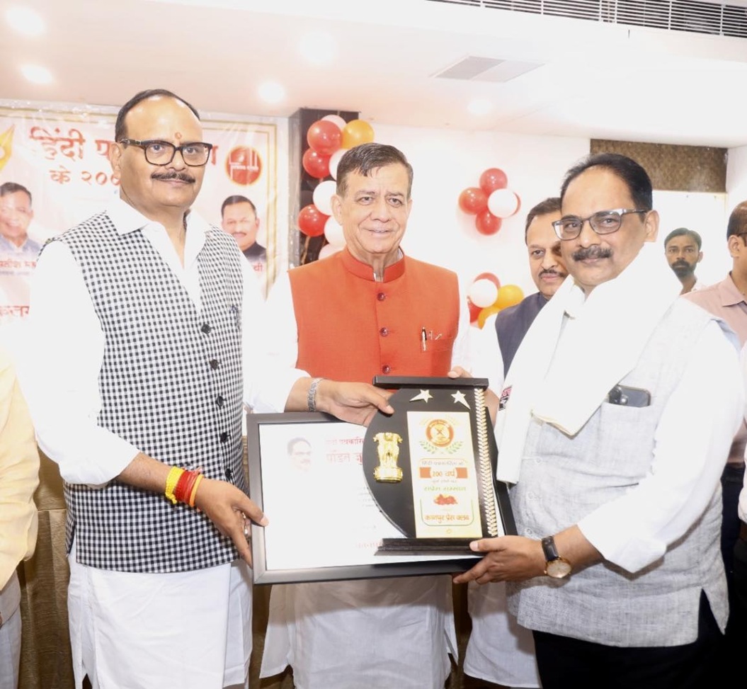 Three men in traditional Indian attire at an award ceremony; the man in the center hands a framed trophy to the man on the right as others look on, with red and white balloons in the background.