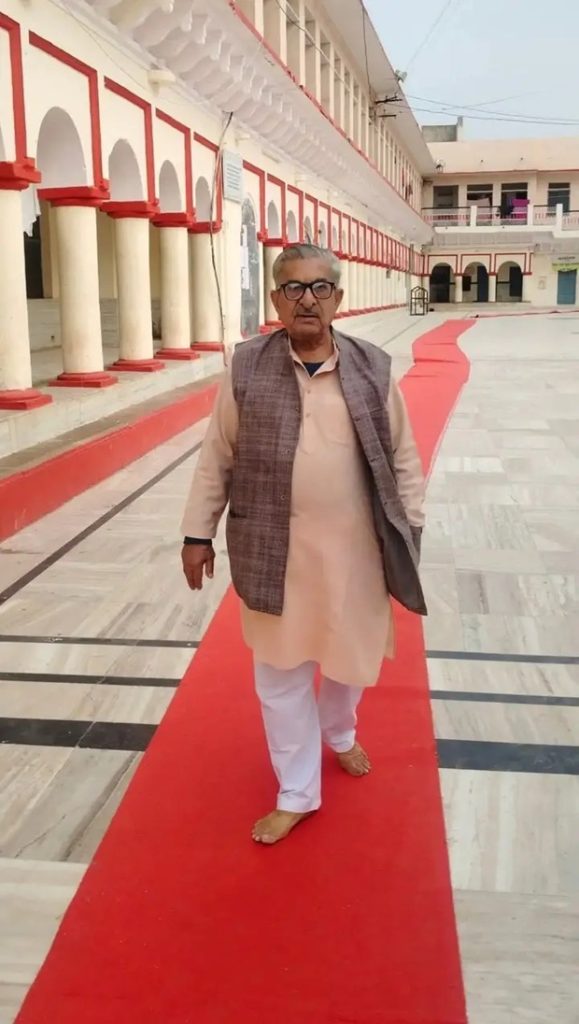 Older man in beige kurta and vest, barefoot, walking on a red carpet along a marble corridor with arches.