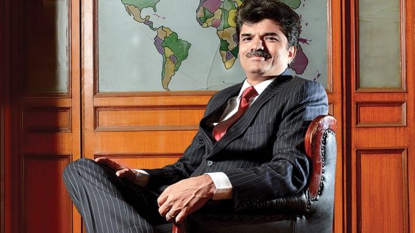 Man in a dark pinstripe suit sits in a leather chair, with a world map on a wooden wall behind him.