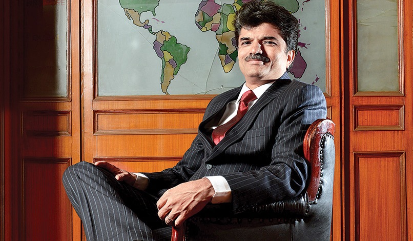 Man in a dark pinstripe suit sits in a leather chair, with a world map on a wooden wall behind him.