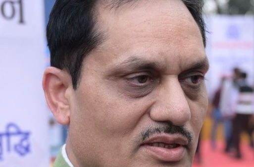 Close-up of a man in a green kurta speaking into microphones at an outdoor event with banners in the background