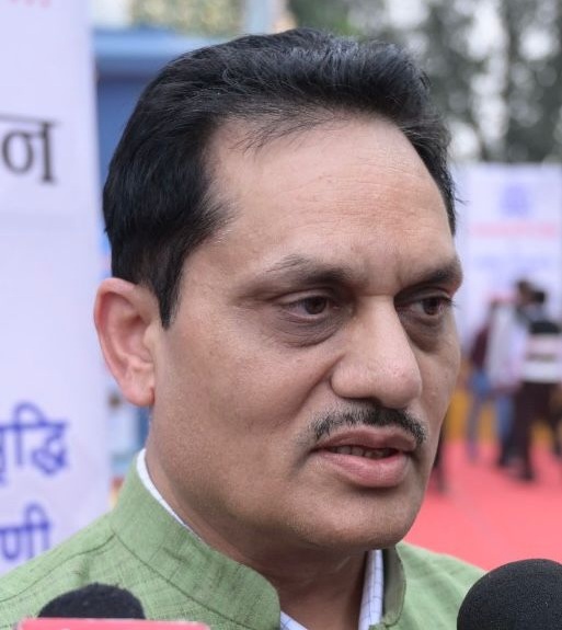Close-up of a man in a green kurta speaking into microphones at an outdoor event with banners in the background
