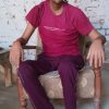 Adult man seated in a worn armchair outdoors, wearing a magenta T‑shirt and purple pants, with a nasal oxygen tube.