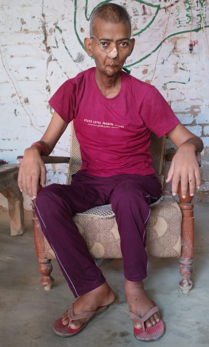 Adult man seated in a worn armchair outdoors, wearing a magenta T‑shirt and purple pants, with a nasal oxygen tube.