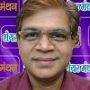 Portrait of a man wearing glasses and a maroon shirt, smiling, against a purple backdrop with Hindi text.