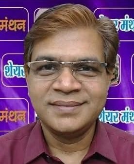 Portrait of a man wearing glasses and a maroon shirt, smiling, against a purple backdrop with Hindi text.