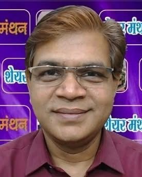 Portrait of a man wearing glasses and a maroon shirt, smiling, against a purple backdrop with Hindi text.