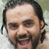 Close-up of a man with dark hair and a beard emerging from water, smiling with his mouth open.