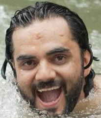Close-up of a man with dark hair and a beard emerging from water, smiling with his mouth open.