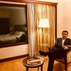 Man in a suit sits in a hotel chair with legs crossed, a lamp beside him and a window showing a bed in the hotel room behind.