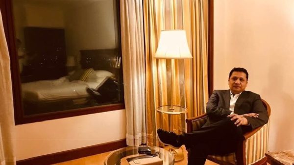 Man in a suit sits in a hotel chair with legs crossed, a lamp beside him and a window showing a bed in the hotel room behind.