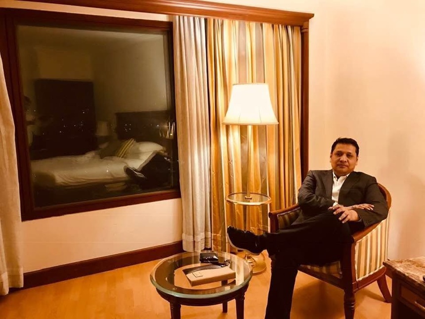 Man in a suit sits in a hotel chair with legs crossed, a lamp beside him and a window showing a bed in the hotel room behind.