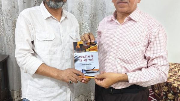 Two men indoors exchanging a book; one wears a white shirt, the other a pink checkered shirt, both smiling.