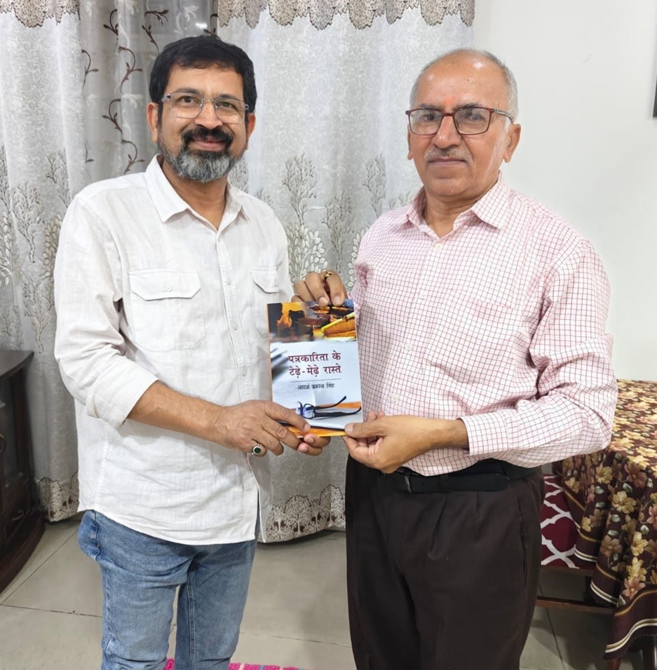 Two men indoors exchanging a book; one wears a white shirt, the other a pink checkered shirt, both smiling.