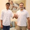 Two men in white shirts shake hands in a living room, with a Gandhi portrait on the wall behind them.