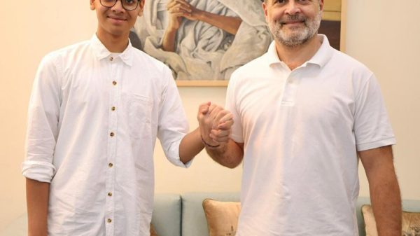 Two men in white shirts shake hands in a living room, with a Gandhi portrait on the wall behind them.