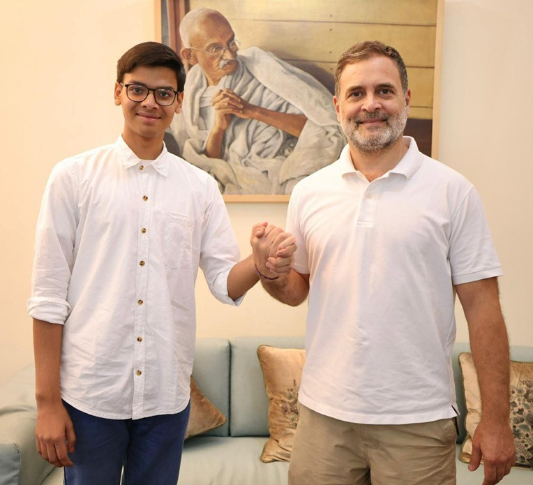 Two men in white shirts shake hands in a living room, with a Gandhi portrait on the wall behind them.