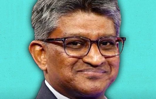 Portrait of a man in a dark suit and glasses, smiling against a teal background.