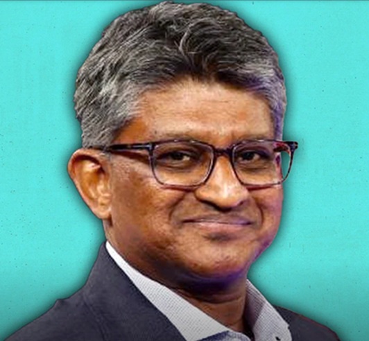 Portrait of a man in a dark suit and glasses, smiling against a teal background.