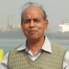 Older man with a red tilak on his forehead, wearing a light striped shirt and beige vest, standing near a harbor with ships in the background.