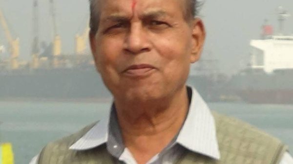 Older man with a red tilak on his forehead, wearing a light striped shirt and beige vest, standing near a harbor with ships in the background.